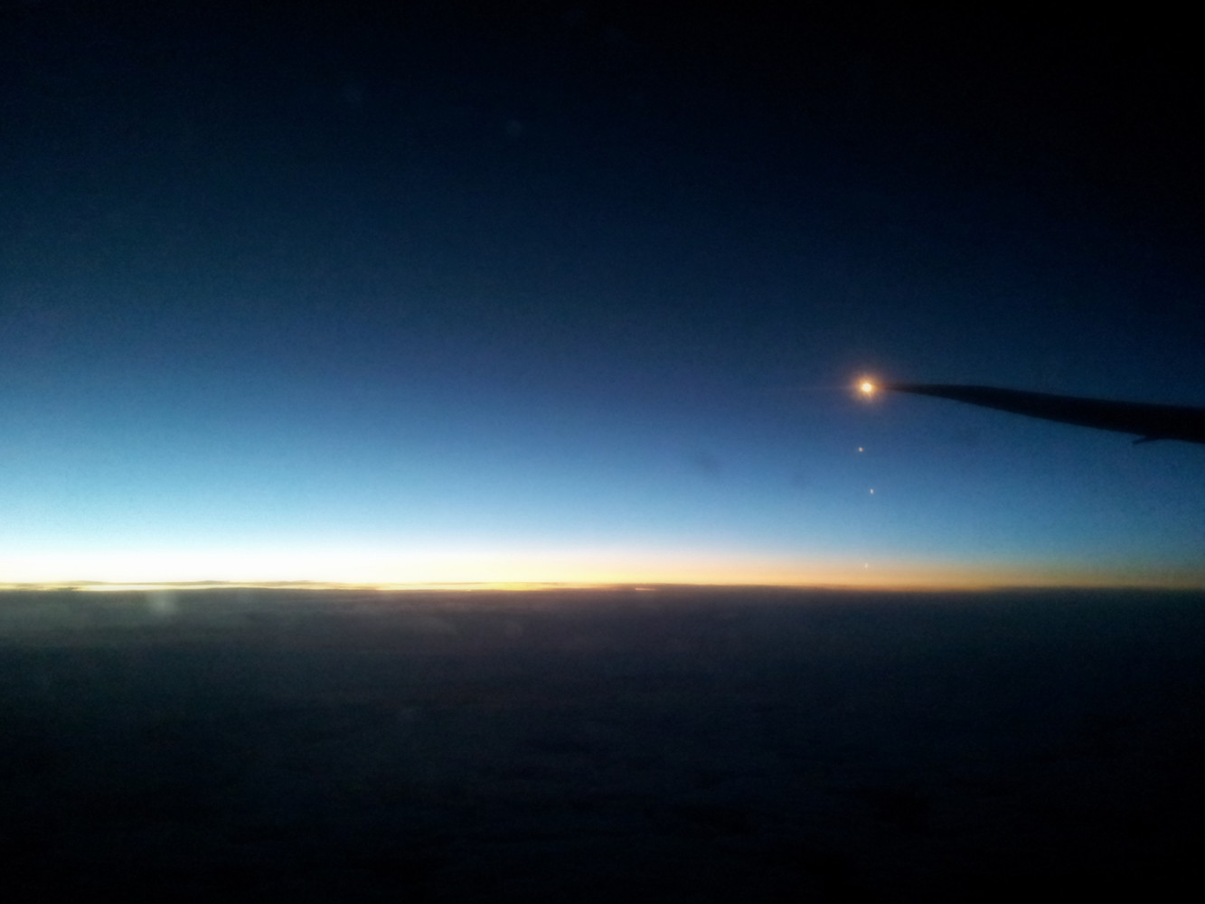Silhouette of the wing of an airplane against a dimly lit horizon at dawn as seen from the passenger seat of an airplane