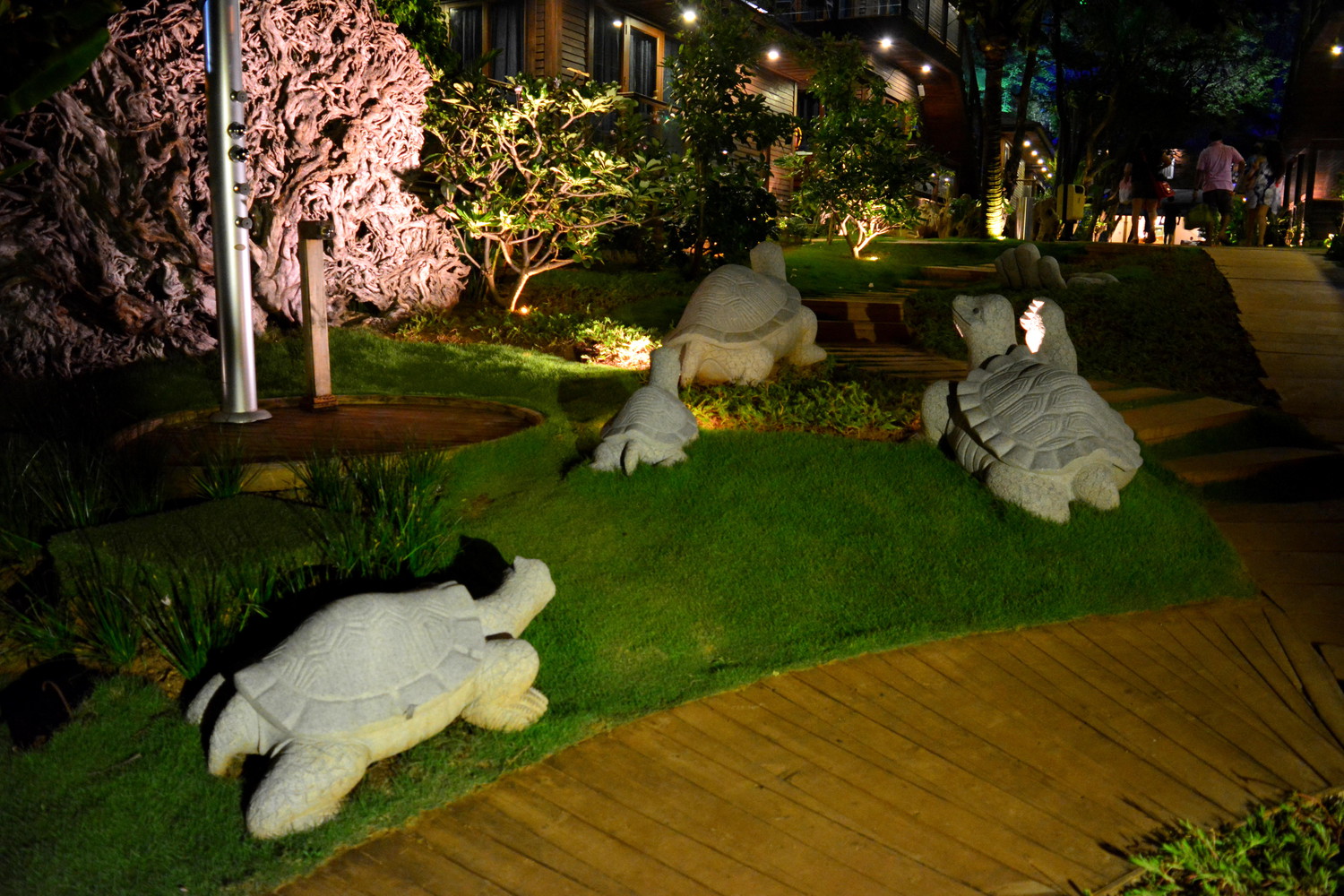 Tortoise sculptures made of stone on green lawn, wooden pavement, and wooden hotel rooms in a resort lit with orange lamps at night