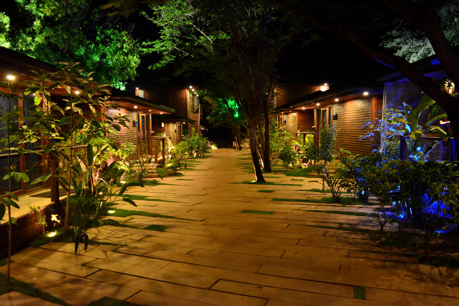 Two rows of wooden hotel rooms with a wooden pavement in between in a resort lit with orange lamps at night