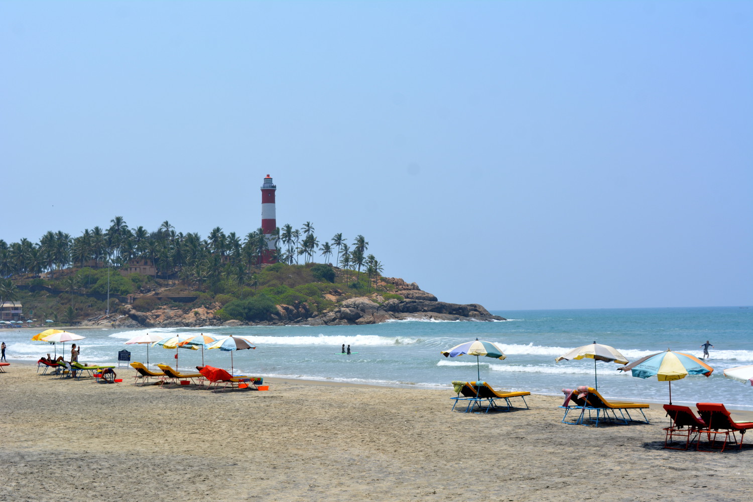 A crescent shaped beach lined with deckchairs and beach umbrellas, and a lighthouse surrounded by coconut palm trees on a hillock