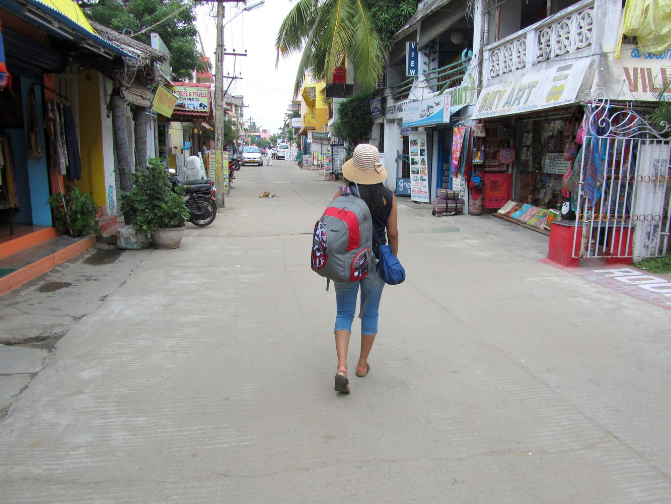 A female traveller with a hat, a backpack, and a purse walking on a street with houses and shops on both sides