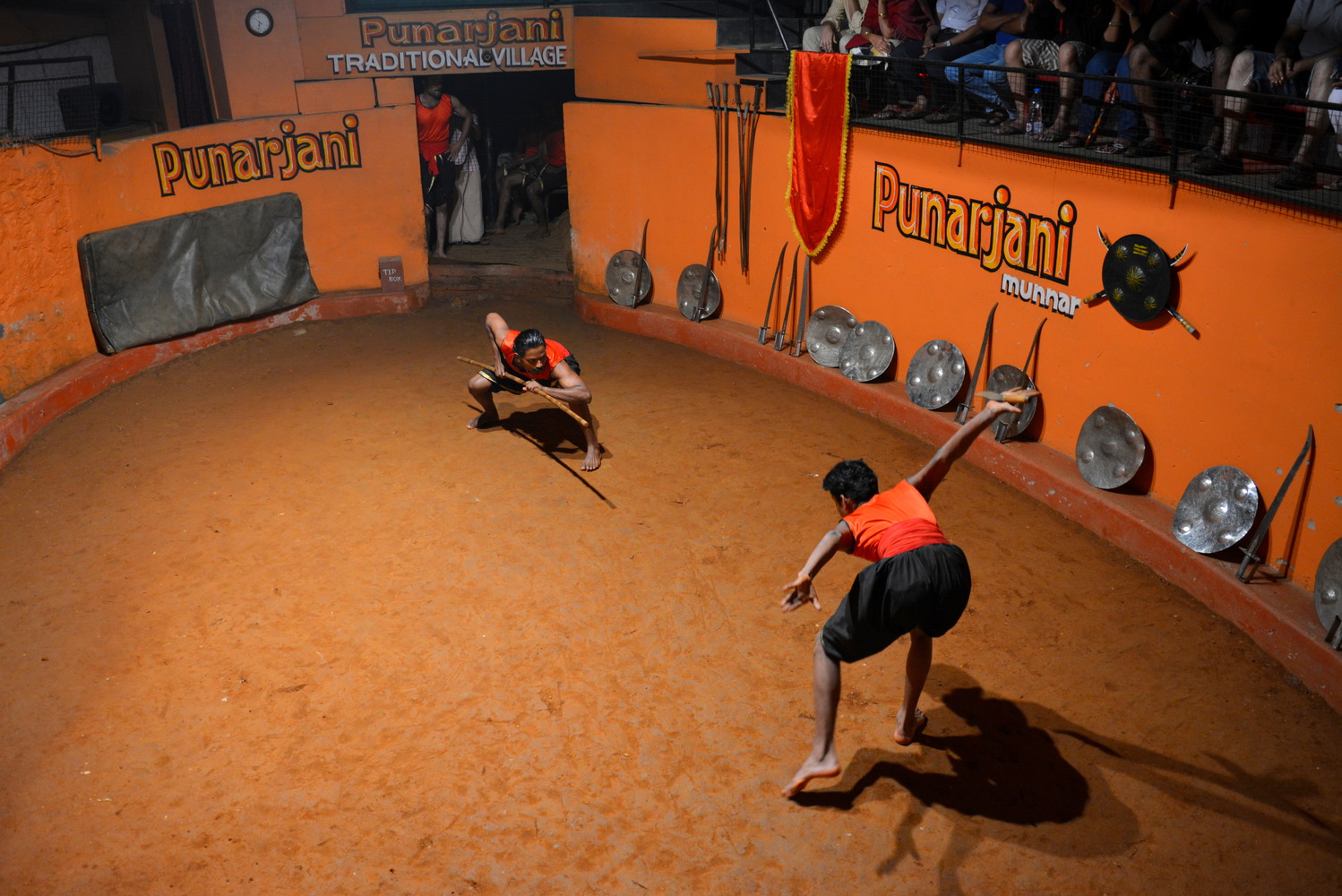 Two martial artists performing in an arena: One artist on the right side is crouching with a stick held in his both hands ready to attack the other artist on the left side while the other artist is looking at him with his hands extended outwards anticipating the move of the first artist