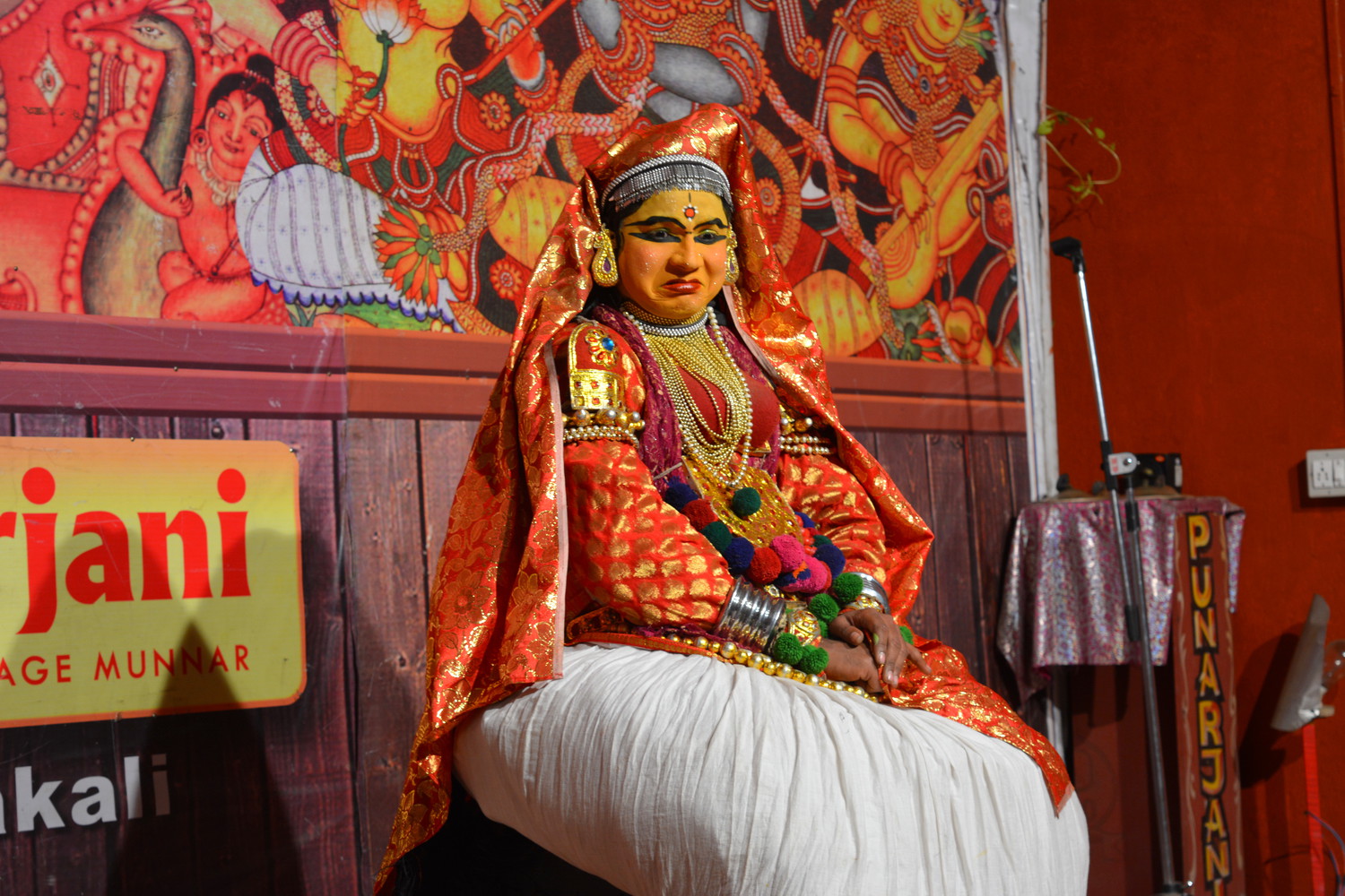 A female Kathakali artist in red costume demonstrating disgust with her facial expressions while seated on a small stool
