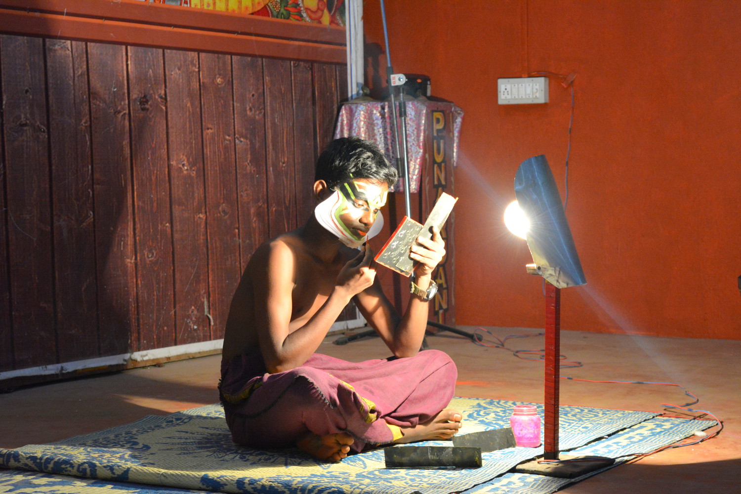 A young boy who is a Kathakali artist applying makeup on his vividly painted face with a small mirror and a piece of paper in his hand, seated on the floor with an electric lamp in front of him