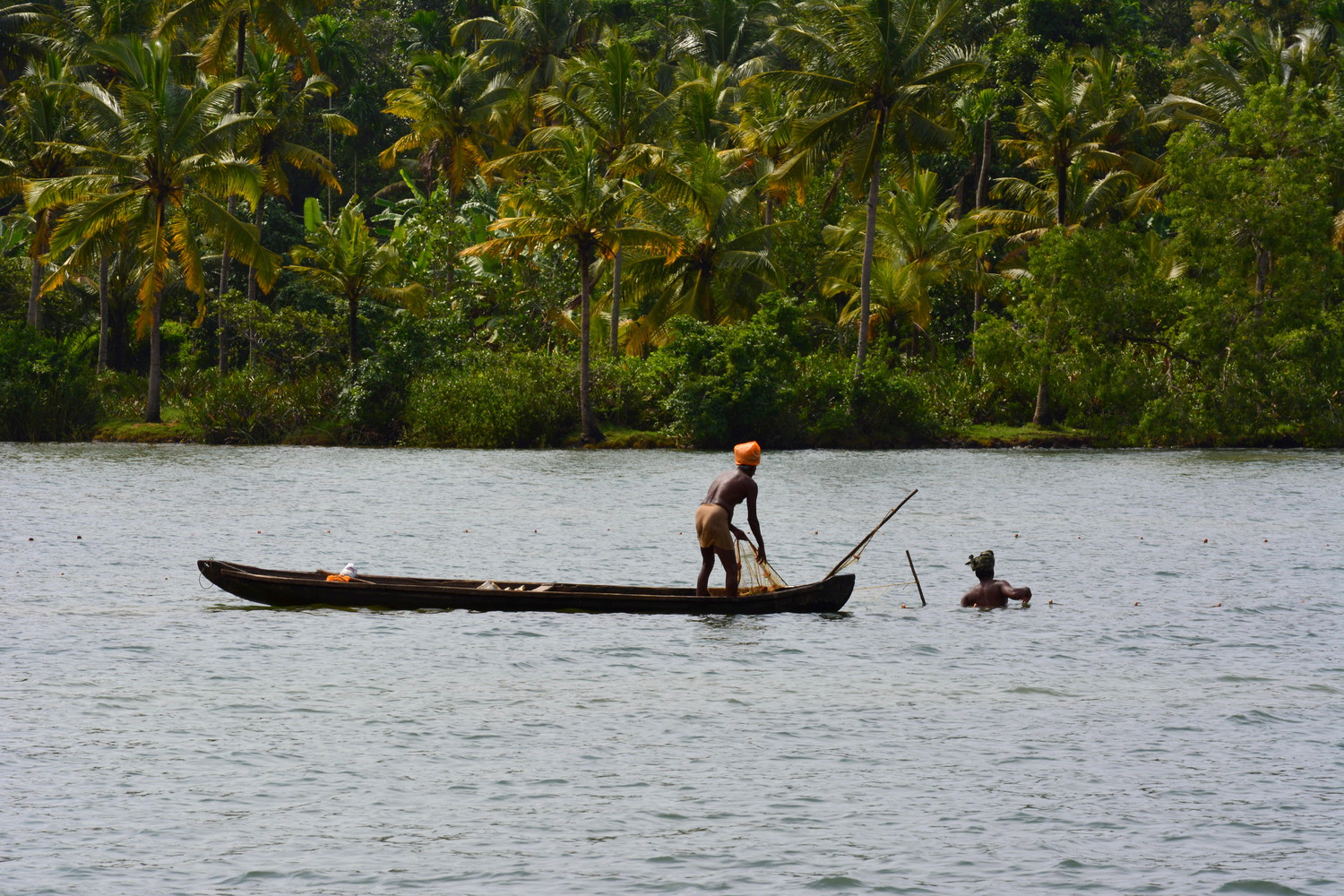 Two fishermen at work in a lake: one standing on a boat pulling a fishing net and the other in water with his body partially submerged
